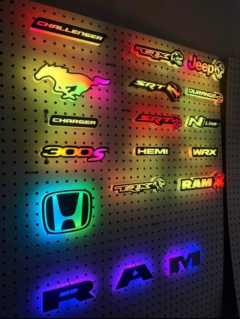 Collection of neon car brand logos on a pegboard wall, featuring Challenger, Charger, Jeep, SRT, Durango, 300S, Hemi, WRX, RAM, Trax, Honda, and BMW in various bright colors